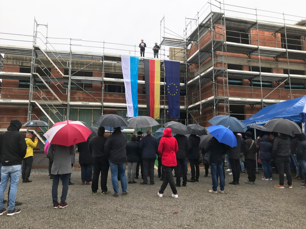 Menschen in Jacken und mit Regenschirmen stehen vor dem Neubau, der noch eingerüstet ist.